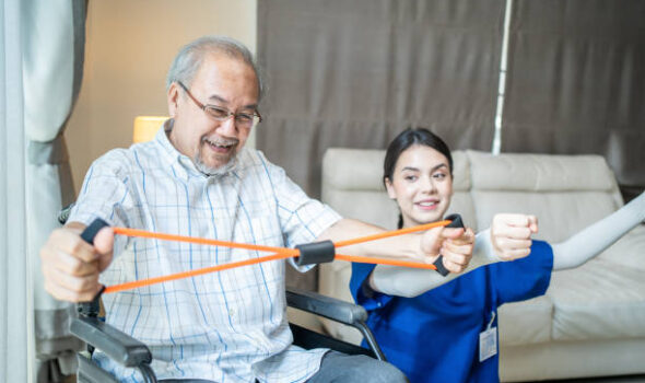 Asian Disabled senior elderly man on wheelchair doing physiotherapist with support from therapist nurse. Older handicapped man using resistance stretch band exercise for patient in home nursing care